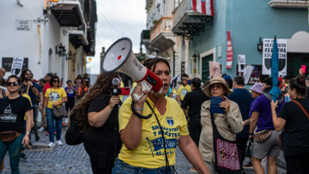 Manifestación de la Federación de Maestros de PR en la Calle Resistencia en el Viejo San Juan. En la imagen se enfoca a una mujer con megáfono alrededor de un gentío.